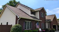 Two-story suburban house with brick and siding exterior, American flag, and neatly trimmed bushes under a clear sky.