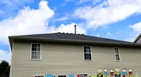 Side view of a beige house with a grey roof under blue sky and a Roof It Forward sign in front.