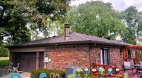 Brick house with gray roof and outdoor seating surrounded by lush green trees under blue sky.