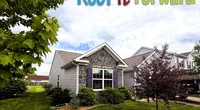 Small house with stone and siding exterior, well-kept lawn, trees, and Roof It Forward logo in sky.