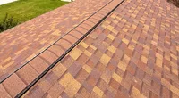 Brown and tan asphalt shingle roof with a ridge vent on a sunny suburban home with grass and cars in background
