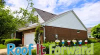 Residential brick and siding house with a pitched roof and Roof It Forward sign in yard under blue sky.