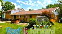 Sunny backyard view of a brick house with patio, garden, and Roof It Forward logo with cartoon children.