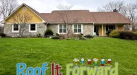 Stone and yellow siding house with green lawn and Roof It Forward colorful campaign sign on front yard.