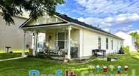 Small beige house with front porch in green yard under blue sky and Roof It Forward logo at bottom.