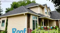 Modern beige two-story house with gray roof surrounded by greenery and the text Roof It Forward overlay.