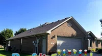 Brick house with gray roof under clear blue sky and colorful Roof It Forward logo at the bottom.