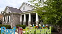 Modern stone and siding house with front porch and Roof It Forward colorful logo in foreground