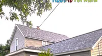 Grey asphalt shingle roofs on a two-story house and garage with a wooden fence in front under tree branches.