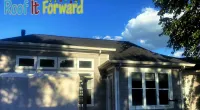Residential house exterior under clear blue sky with Roof It Forward logo in top left corner