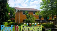 Brick house with green lawn and trees under a partly cloudy sky with Roof It Forward logo and children graphics.