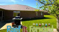 Backyard view of a brick house with a grill and 'Roof It Forward' text with cartoon kids overlay.