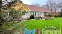 Stone house with yellow siding and brown roof surrounded by greenery and Roof It Forward logo at bottom