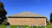 Brick house with a large roof and colorful Roof It Forward logo on green lawn under a clear blue sky.