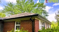 Brick house with a newly installed roof and gutters under a bright blue sky with green trees and bushes.