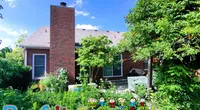 Backyard view of brick house with chimney surrounded by lush greenery under a sunny blue sky.