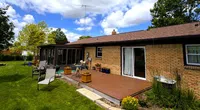 Backyard view of a brick house with a wooden deck, patio furniture, green lawn, and partly cloudy sky above.