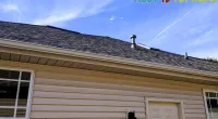 Residential house exterior showing beige siding, shingled roof with vents, windows, and a white door under blue sky.