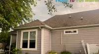 Suburban house exterior with beige siding, a gray roof, windows, plants in yellow pots, and a Roof It Forward logo.