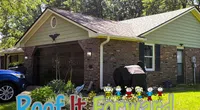 Suburban brick house with a dark garage door, garden, covered barbecue grill, and a blue car parked nearby.