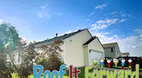 Sunny suburban house with beige siding and a green lawn under a blue sky and the Roof It Forward logo.