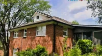 Brick house with a shingled roof surrounded by green trees and bushes under a partly cloudy sky.