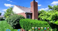 Red brick house with lush green bushes under a bright blue sky, featuring Roof It Forward text with cartoon kids.