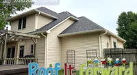 Two-story beige house with dark gray roof and Roof It Forward charity logo overlay in backyard.