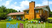 Suburban brick house with lush green lawn and Roof It Forward logo featuring colorful children graphics.