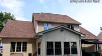 Modern beige house with new brown roof under blue sky and Roof It Forward charity logo in corner.