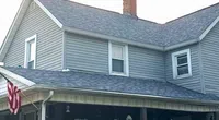 Two-story gray house with new shingled roof, a brick chimney, American flag, and green bushes in front yard.