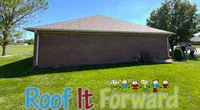 Sunny backyard with brick house wall and Roof It Forward colorful sign on green grass under trees.
