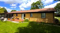 Single-story brick house with a large green lawn under a partly cloudy blue sky, captioned Roof It Forward.