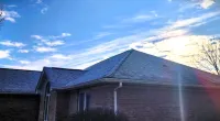 Brick house with sloped roof under a blue sky with scattered clouds and sunlight flare.