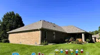 Brick house with a well-maintained lawn under a clear blue sky, featuring Roof It Forward logo with colorful kids.