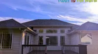 Modern house exterior with dark roof shingles, large windows, and wooden deck under blue sky with clouds