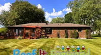 Single-story brick house with red deck and pergola, surrounded by green lawn and large trees under a blue sky.