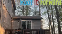 Gray house with wooden deck and patio furniture under Roof It Forward logo with cartoon children and colorful text.