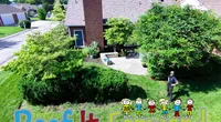 Green suburban backyard with trees, a brick chimney house, and a man standing near a colorful Roof It Forward sign.