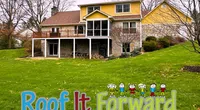 Yellow and stone house with large backyard and colorful Roof It Forward logo on green grass in front yard.