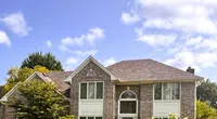 Large two-story brick house with arched front door, multiple windows, surrounded by green trees and shrubs under blue sky
