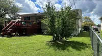 Single-story house with wooden deck, lush green yard, tree in front, chain-link fence, and blue sky with clouds.