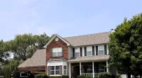 Two-story brick and siding suburban house with a well-maintained lawn, trees, and a clear sky overhead.