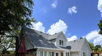 Sunny backyard patio with outdoor furniture, a white house with a gray shingled roof, and a clear blue sky.