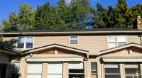 Two-story beige house with dark roof under blue sky and tall green trees in the background