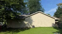 Side view of a beige house with a neatly mowed lawn and large trees under a clear blue sky