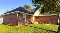 Brick house with a dark shingled roof under clear blue sky and a large green tree on the lawn
