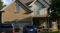 Two-story suburban house with brick and siding exterior, garage, driveway with two parked vehicles, and green lawn under blue sky