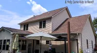 Two-story beige house with new brown roof, patio umbrella, outdoor seating, and Roof It Forward charity logo.