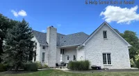 Large white brick house with gray roof under blue sky and Roof It Forward charity logo in corner.
